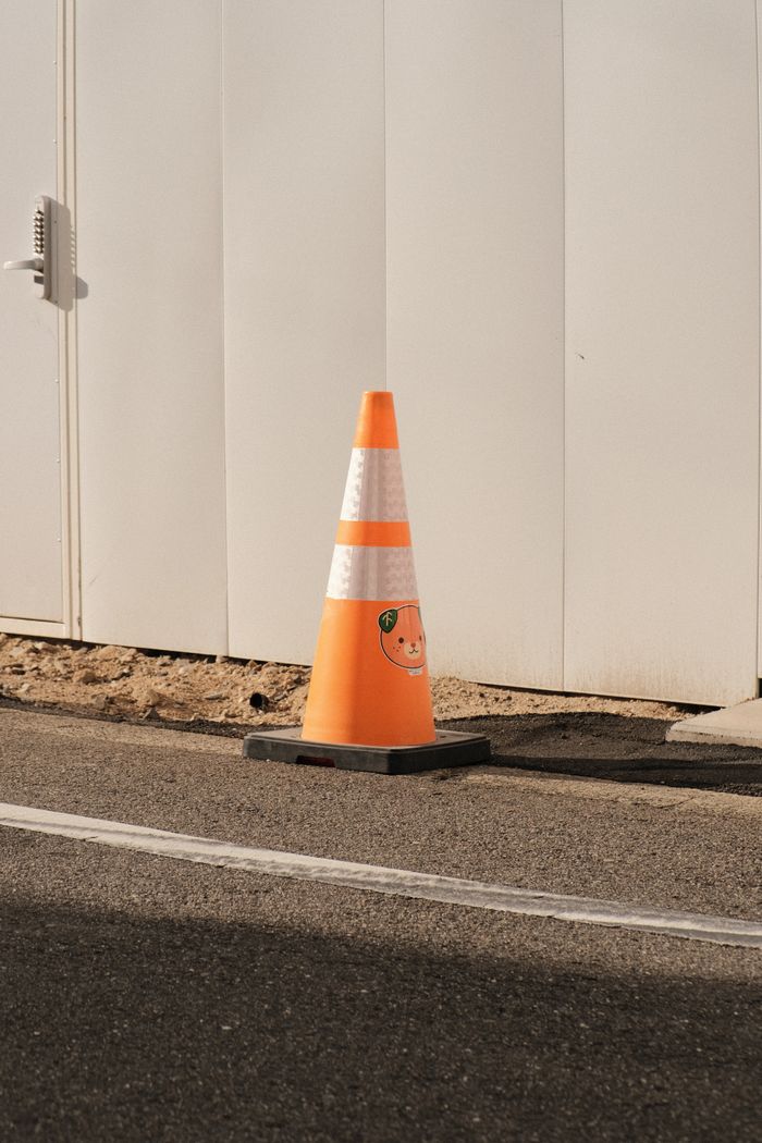 A brightly colored traffic cone placed on a city street beside a white wall.